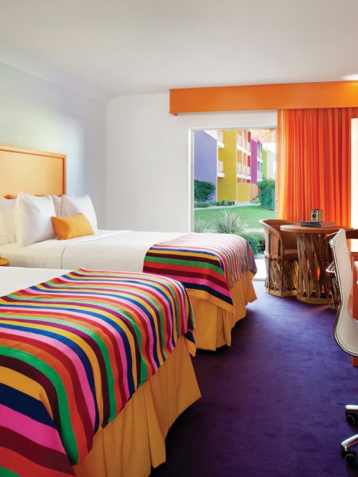 Colorful hotel room with two beds, desk with rolling chairs, and flat TV