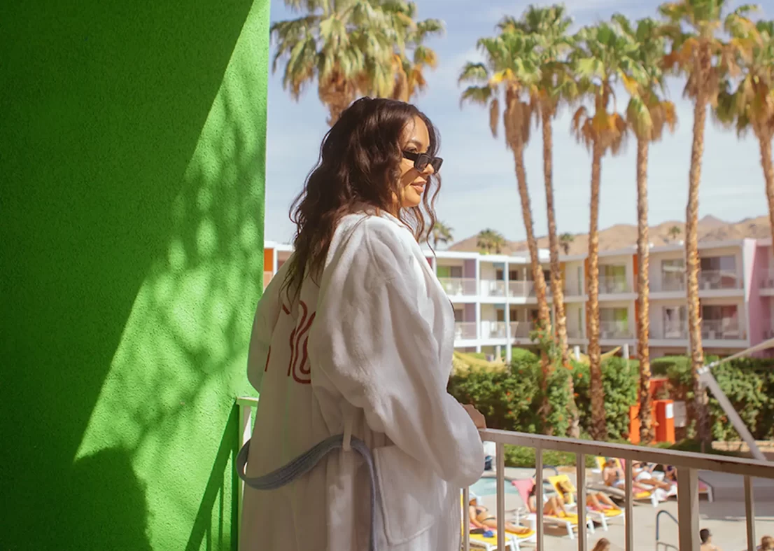 Woman in a white robe on a balcony overlooking a pool area at a Palm Springs hotel with palm trees.
