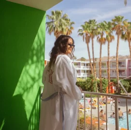 Woman in a white robe on a balcony overlooking a pool area at a Palm Springs hotel with palm trees.