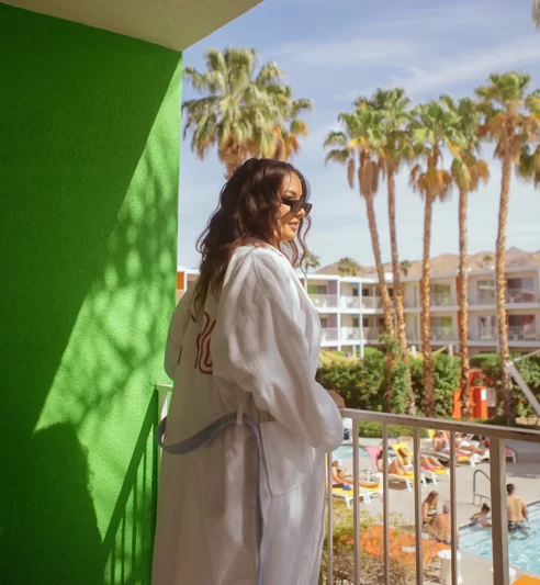 Woman in a white robe on a balcony overlooking a pool area at a Palm Springs hotel with palm trees.