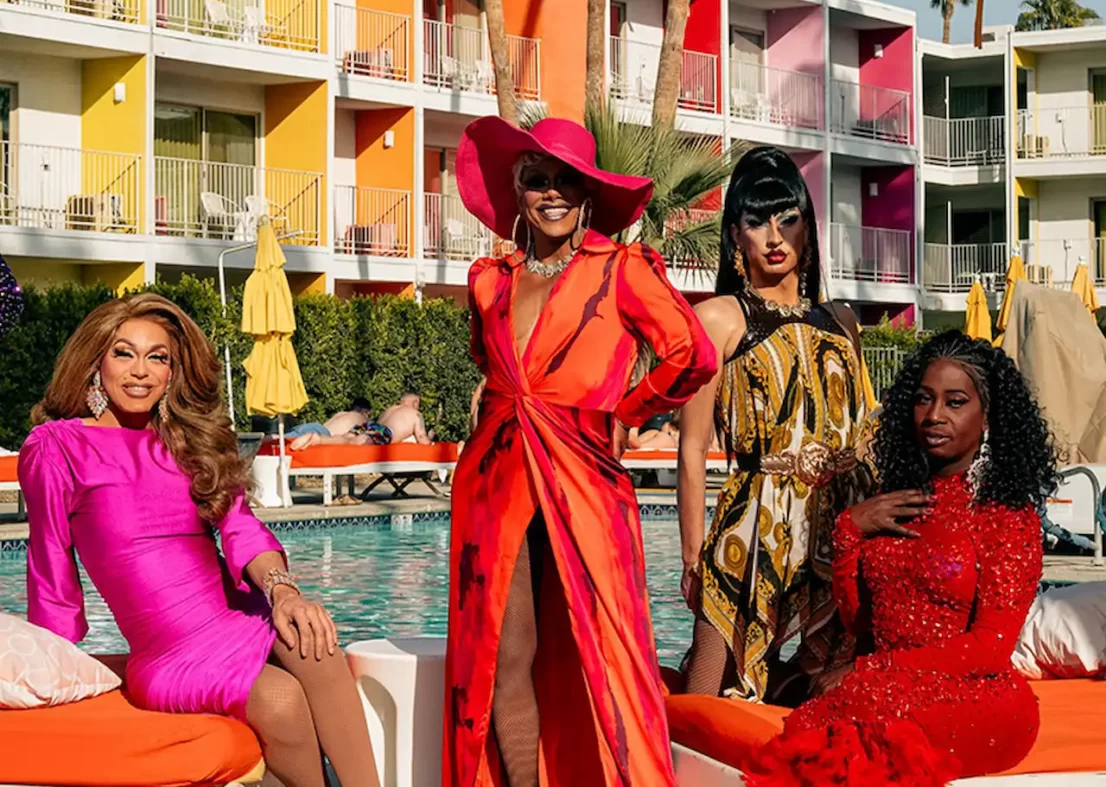 Drag performers in bold red and pink outfits relaxing poolside at a colorful hotel.