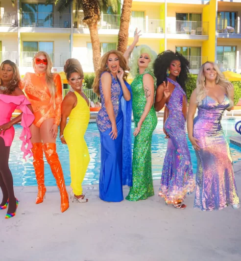 Seven people in vibrant colorful outfits posing in front of a pool and a hotel building.